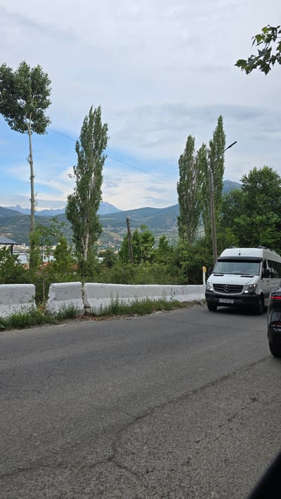 The image shows a white Mercedes-Benz Sprinter van driving on a paved road in Charvak, Uzbekistan. The van is white with tinted windows and has the license plate AZ 683ZC. In the foreground, there is a concrete barrier along the side of the road, and a dark-colored vehicle is partially visible on the right. In the background, there are tall, slender trees, green foliage, and mountains in the distance under a cloudy sky. The scene appears to be a roadside view on a cloudy day.