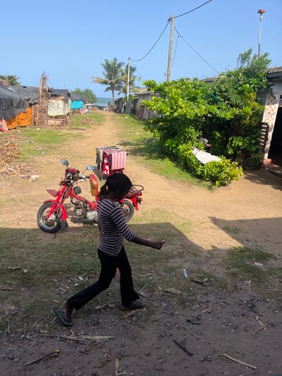 In Moratuwa, Sri Lanka, a young girl with dark hair, wearing a striped long-sleeved shirt and dark pants, walks along a dirt path lined with simple dwellings. To her left, a bright red motorcycle is parked. Behind the motorcycle, a clothes drying rack holds colorful folded fabrics. Palm trees and lush green foliage are visible in the background, with a glimpse of the ocean beyond. The environment appears to be a coastal community, with some structures showing signs of temporary construction and the ground littered with debris. The lighting suggests a sunny day.