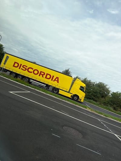A yellow articulated lorry with "DISCORDIA" written in red lettering on its side is driving on a paved road. The truck is mostly visible, with its cab and trailer occupying a significant portion of the image. The truck is positioned at an angle, moving away from the viewer and towards the right side of the frame. The road surface is asphalt, with white lane markings visible. To the left of the road, there is a grassy verge with some foliage and trees. The sky is overcast with grey clouds. The scene appears to be located in a rural or semi-rural area, possibly on a highway or main road. The image is taken from a slightly elevated perspective, looking down at the truck and road. No people are clearly visible.