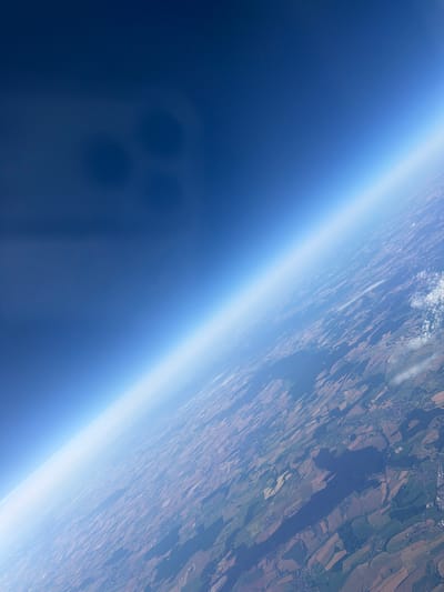 The image is an aerial view looking down at the earth's atmosphere and landscape from a high altitude. The upper portion of the image is dominated by a deep blue sky, transitioning to a lighter blue and then to a hazy white band at the horizon, representing the curvature of the Earth and atmospheric layers. Below this atmospheric layer, the landscape is visible, appearing as a patchwork of fields with varying colors of brown, green, and tan. Some areas show signs of human habitation, with visible towns and roads. There are also some scattered white clouds in the lower right quadrant. In the upper left portion of the image, there are four dark, circular, blurry shapes that appear to be reflections or shadows on the camera lens or window. The location is identified as the city of Werl, Germany, which can be inferred from the details of the landscape showing agricultural fields and urban development consistent with that region.
