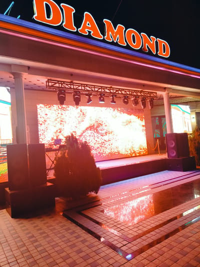 An outdoor night scene in Aksu, Kazakhstan, features the illuminated sign "DIAMOND" in large orange letters with white outlines, set against a dark sky. Below the sign, a stage is equipped with a large LED screen displaying a bright, abstract pattern of white and orange light, reminiscent of fireworks or sparks. Above the screen, a truss structure holds several spotlights. Two large black speakers are positioned on the left side of the stage, and another pair is on the right, near a golden rope barrier. In front of the stage, a tiered tiled area reflects the stage lighting and the LED screen's display. A small, dark green shrub sits in the foreground, partially obscuring the stage. The overall impression is of an entertainment venue prepared for an event.