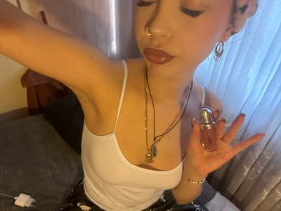 An individual is depicted in an indoor setting, identified as Haguenau, France. The subject, a female, is shown from the upper torso, with her left arm raised. She is holding a small, silver-capped cosmetic bottle, containing a reddish-brown liquid and possibly branded "KIKO," in her left hand. Her facial features include a nose piercing on her right nostril, glossy reddish-brown lips, and a dangle earring in her right ear. Her attire consists of a white spaghetti-strap tank top and dark, patterned lower garments. Light hair growth is discernible in her raised left armpit. She wears a gold-colored necklace with multiple pendants, one resembling a Buddha figure, and a multi-colored beaded bracelet on her left wrist. The background includes light-colored curtains, a dark grey fabric surface possibly indicative of bedding, and a dark brown furniture item. A white wall charger is resting on the dark grey fabric surface.