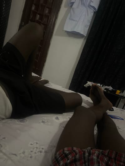 The image shows a close-up view of a person's legs and feet, resting on a patterned bedsheet. The person is wearing black shorts and a red and black checkered garment on their right leg. The setting appears to be indoors, possibly a bedroom, with a dark curtain visible in the background and some white clothing draped over a surface on the left side of the frame. The lighting is dim, suggesting it could be evening or night. There is no visible text in the image. The focus is on the lower body of the individual, with the legs crossed.