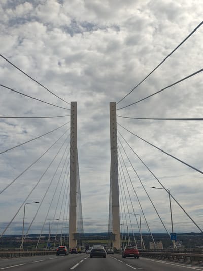 This image shows a view from the M25 motorway crossing the Queen Elizabeth II Bridge in Dartford, United Kingdom. The perspective is from within a vehicle, looking forward. Two prominent white cable-stayed bridge towers dominate the center of the frame, with numerous cables extending upwards and outwards, creating a symmetrical pattern against a cloudy sky. Several vehicles are visible on the multi-lane carriageway, moving away from the viewer. The bridge deck, with its concrete barriers and lampposts, is also clearly depicted. In the distance, a landscape of rolling hills and scattered buildings can be seen. The overall atmosphere is overcast, with diffused light.