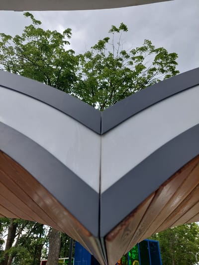 The image shows a close-up view of the underside of a V-shaped modern roof structure, possibly part of a gazebo or pavilion, with trees and a cloudy sky visible in the background. The roof is constructed with alternating bands of light grey material and dark grey trim, meeting at a central V-shaped seam. Below this, wooden planks are visible, running horizontally. In the background, through the gap in the roof structure, lush green trees with dense foliage are seen against a pale, overcast sky. The scene appears to be outdoors, in a park or recreational area, given the presence of trees and what looks like some colorful playground equipment or art installations in the lower right portion of the image. The location is identified as Alikonovka, Russia. There are no people or animals visible in the image.