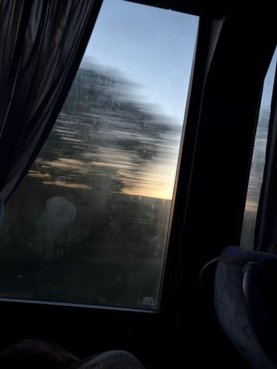 The image depicts a view through a bus window at dusk, with dark curtains on the left side partially obscuring the scene. The window is smudged, and the exterior view shows a blurred landscape with streaks of light, suggesting movement. The sky is a pale blue with hints of orange and yellow near the horizon, indicating sunset. There are dark, indistinct shapes in the middle ground, possibly trees or buildings. In the foreground, on the right, a portion of a blue and grey upholstered seat is visible, suggesting the perspective is from inside a vehicle. The overall impression is of a journey in progress, with a melancholic or peaceful ambiance due to the low light and blurred scenery. The location in Stoicani, Moldova, is not evident from the visual cues within the image itself.