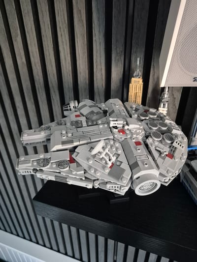 The image shows a LEGO model of the Millennium Falcon spaceship displayed on a black shelf in what appears to be a home or office setting. The background consists of dark, vertically striped wooden panels. To the right of the Millennium Falcon, a white speaker is visible, and in the background, a small LEGO model of the Empire State Building is also present. The lighting is soft, casting some shadows. The scene does not provide any information about Barbing, Germany.