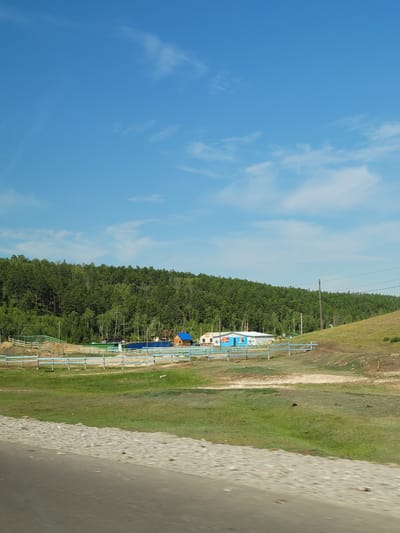 The image depicts a rural landscape under a clear blue sky with scattered white clouds. In the foreground, a dirt road is visible, with a blurred grassy field stretching beyond it. A low, blue-and-white fence runs horizontally across the middle ground, separating the field from a gently sloping hillside.

On the hillside, there are several buildings. One prominent building is a small, wooden house with a bright blue roof. Adjacent to it are other structures, including a long, white building with blue trim and some smaller, colorful buildings. These structures appear to be part of a rural settlement or farm.

Behind the buildings, a dense, green forest covers the hillside, extending to the horizon. The trees are lush and appear to be deciduous. Power lines run across the scene, supported by wooden poles. There are no people or animals clearly visible in the image. The overall impression is one of a peaceful, natural environment with a small human presence. The location is identified as Karapatskoye, Russia.