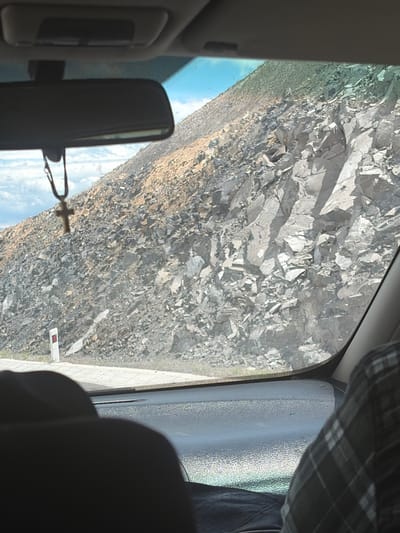 The view from inside a vehicle shows a steep, rocky hillside alongside a road. The hillside is composed of light-colored rocks and scree, and appears arid. The road is visible as a narrow strip along the base of the hillside. The rearview mirror and part of the vehicle's interior are in the foreground. A plaid shirt is visible at the top right of the frame. A hanging cross decoration is attached to the rearview mirror. A road marker is visible along the road. The location is identified as Agarak, Armenia.