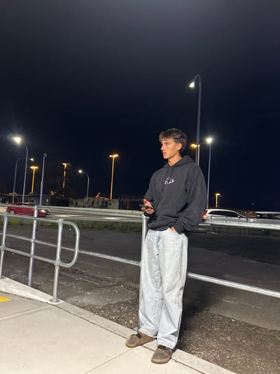 A full-body, low-angle shot captures a young male standing near a metal railing at night. He is wearing a black hoodie, light-wash baggy jeans, and brown Birkenstock sandals. The man is holding a black smartphone in his right hand and looking to his left. Behind him, several tall streetlights illuminate the scene. In the background, a red car is visible on a road, and beyond that, a dark landscape with some structures and more streetlights. The ground beneath the man is concrete, with a darker, rougher surface behind the railing. The overall atmosphere is dimly lit, typical of an urban setting at night. The location is identified as Beaconsfield, Australia.