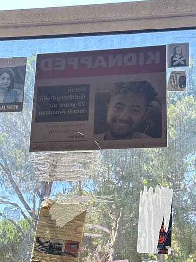 This media file captures a close-up view of a window pane covered with various notices and torn pieces of paper. The dominant subject is a "KIDNAPPED" poster featuring a man's face, appearing to be a missing person flyer. The text on the poster is partially visible and appears to be in English and possibly another language, with the word "KIDNAPPED" written vertically.
Scattered around the main poster are smaller, abstract images or icons, one of which resembles a stylized silhouette of a person. Other pieces of paper are heavily torn and weathered, with some exhibiting what looks like handwritten text or printed graphics in a foreign script, possibly Arabic given the location context. One particularly torn piece has a fragment of what appears to be a barcode or serial number.
The background visible through the window suggests an outdoor scene with green foliage and trees, indicating it is daytime. The quality of light and the dappled shadows imply sunlight filtering through leaves. The overall impression is one of urgency and perhaps desperation, given the "KIDNAPPED" poster and the distressed state of some of the other papers. The window itself appears to be part of a building, with a textured frame visible on the left side. The presence of multiple, overlapping notices suggests a public space or a location where information is frequently posted and updated.