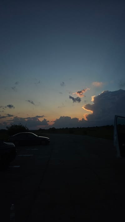 The image captures a twilight scene in Buhovtsi, Bulgaria. The sky dominates the upper two-thirds of the frame, transitioning from a deep blue-grey at the top to lighter blue and then to warm orange and yellow hues along the horizon, indicative of sunset. Numerous clouds are present; scattered, darker clouds are visible across the mid-sky, while a large, dense cloud formation on the far right is intensely backlit by the low sun, creating a dramatic, jagged outline of bright orange and white light. The lower third of the image is significantly underexposed, rendering the foreground in deep shadow. This foreground consists of an asphalt surface, consistent with a road or parking lot, where faint white line markings are discernible on the left side. A dark-colored sedan, appearing to be a BMW E90 series, is parked on the left, facing right, its silhouette sharply defined against the slightly brighter horizon. A partial view of another dark vehicle is visible on the extreme lower-left edge. On the far right, the open door of a vehicle, possibly a van or SUV, is partially visible, with its interior and structural elements appearing as dark silhouettes. A light-colored plastic bottle is present on the ground in the lower-left corner of the frame. No individuals are visible.