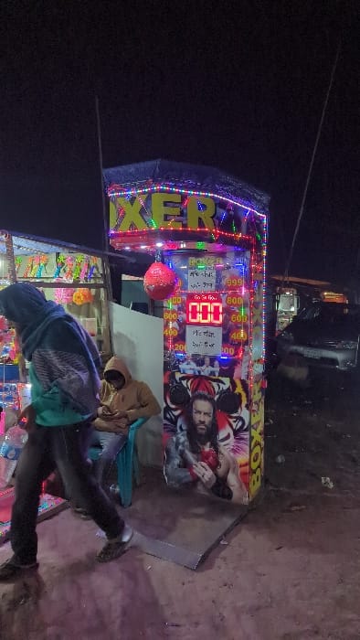 A "BOXER" arcade machine with flashing lights displays a score of "000." The machine features images of wrestlers. Two people stand nearby, one wearing a brown jacket and another in a blue hoodie. The surrounding area is dark and appears to be an outdoor setting.