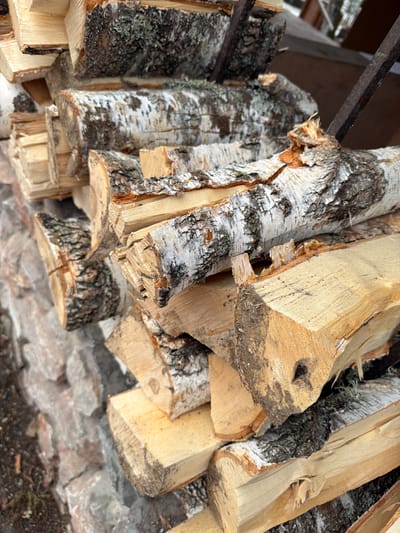 The image shows a stack of chopped firewood, primarily birch, located in Taskino, Russia. The wood is stacked outdoors, seemingly under a partially visible wooden structure. The cut ends of the birch logs show the distinctive white bark and exposed wood grain. The area beneath the firewood appears to be a combination of stone or concrete fragments and exposed soil.