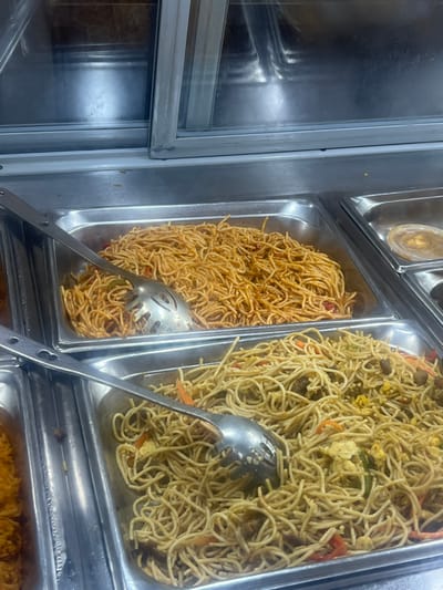 The scene depicts a static, close-up view of a food service display within a commercial establishment in Ikotun, Nigeria. The image focuses on two large, rectangular stainless steel chafing dishes filled with prepared pasta dishes.
The upper chafing dish contains a substantial quantity of red-orange colored spaghetti, uniformly coated in a sauce, with a large, silver-colored metal slotted serving spoon resting within the pasta.
The lower chafing dish holds a volume of light brown, thin noodles, also with a large, silver-colored metal slotted serving spoon embedded. This noodle dish is visibly mixed with various ingredients, including red bell pepper strips, smaller pieces of green vegetables, pale yellow scrambled egg fragments, and small, dark, irregularly shaped protein pieces.
To the upper right, a partial view of another stainless steel tray is visible, containing a small, clear plastic cup with a light brown, viscous substance. The background above the food trays features reflective surfaces, consistent with a display case or sneeze guard, showing blurred reflections of the environment. No individuals are present in the frame, and no active interactions are depicted.
