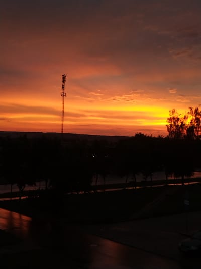 The image captures a dramatic sunset over Vawkavysk, Belarus, casting a vibrant orange and yellow glow across the sky. A tall, dark silhouette of a communication tower stands prominently against the colorful horizon. Below, a dense line of dark trees forms a silhouette, obscuring the details of the landscape beneath. In the foreground, the ground is dark, with a wet sheen suggesting recent rain, reflecting the colors of the sky in streaks of orange and red. A dark-colored car is partially visible on the right side of the frame, parked on what appears to be a street or parking lot. The overall scene is atmospheric, with the setting sun creating a striking contrast between light and shadow.