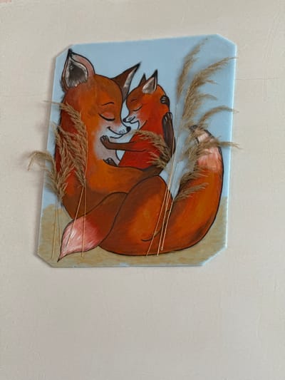 A painting of two foxes, one adult and one cub, embracing each other is displayed on a wall. The adult fox is on the left, with its eyes closed and its head tilted towards the cub. The cub is on the right, nestled into the adult's embrace, also with its eyes closed. Both foxes are depicted with orange fur and black accents on their ears and noses. The background of the painting is a light blue, with a sandy-colored base. Sprigs of dried grass are attached to the painting, framing the foxes. The wall appears to be a neutral, light-colored surface. This image was taken in Tskhinvali, South Ossetia.