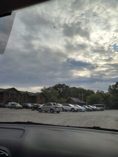 An exterior, wide shot captured from inside a vehicle shows a parking lot with several cars. In the background, a wooden building with a green roof displays the word "HOTEL" in green lettering. Trees line the back of the property. The sky is overcast with scattered sunlight breaking through. The image is located in Shcherbani, Ukraine.