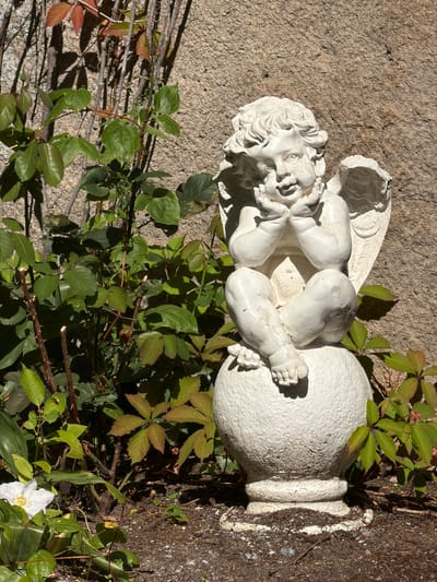 An outdoor close-up shot captures a white cherub statue sitting on a textured, spherical pedestal. The statue, with its head tilted and hands resting on its chin, appears to be made of plaster or concrete and shows signs of wear, with chipped paint. Behind the statue, a rough, textured stone wall serves as the backdrop, with shadows cast across it. To the left and right of the statue, lush green foliage and some reddish-brown leaves are visible, suggesting a garden setting. A single white flower with yellow accents blooms in the lower left corner. The ground is covered in dark soil and small pebbles. The lighting suggests it is daytime, with sunlight creating strong contrasts and shadows. The scene appears to be in a residential or public garden area.