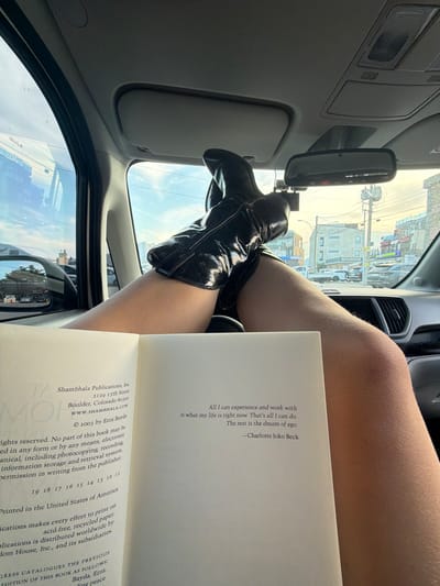 The image shows a view from inside a car. A pair of legs, wearing black patent leather boots, are positioned on the dashboard, partially obscuring the windshield. A book is held in front of the legs, open to a page containing text and publication information about Shambhala Publications, Inc., and a quote by Charlotte Joko Beck. Through the windshield, a street scene is visible, suggesting an urban or suburban environment. Buildings, utility poles, and possibly parked cars can be discerned. The scene is likely in Toseong-myeon, South Korea, based on the location information, as identified on the street sign that says "GAYANG". The rearview mirror is also visible, as well as part of the car's interior, including the sun visor and air vents.