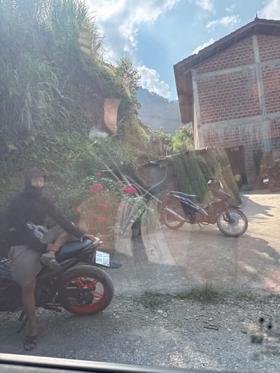 A partially obscured view depicts a scene outside, likely captured through a window. In the foreground, a person in casual clothing (gray shorts, dark shirt) is seated on a black motorcycle with red-rimmed wheels, license plate “25-B7 362.52”. The motorcycle rests on a gravel surface. Further into the scene, additional motorcycles are visible near a building with an unfinished brick facade. Bundles of green reeds lean against the wall beside one of the motorcycles. A verdant hillside, dotted with pink flowers, rises in the background. The image appears to have been taken in the Mậu Duệ Commune of Vietnam due to the presence of the Vietnam license plate on the motorcycle.