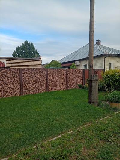 In Shostka, Ukraine, an exterior residential scene unfolds. A well-maintained lawn dominates the foreground, bordered by a concrete strip. Beyond the lawn, a decorative concrete fence with a brown, textured pattern stretches horizontally across the frame. To the left of the fence, stacked lumber is visible, suggesting recent construction or storage.
In the background, a light-colored house with a grey metal roof stands partially obscured by lush green foliage. A wooden utility pole with visible markings, including the numbers "11" and "67," stands prominently in the right foreground, adjacent to the lawn and fence. The sky above is partly cloudy, with patches of blue peeking through. The overall impression is one of a suburban or rural residential setting.