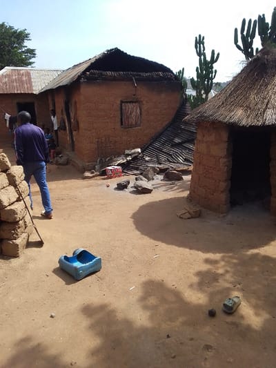 The image shows a scene in Kwal, Nigeria, located at coordinates 9.803 latitude, 8.646 longitude. Two mud brick buildings, both with thatched roofs, are the focus of the scene. The building on the left is partially collapsed, revealing the red brick interior and a metal roof beneath. The thatch roof on the left building is partially burnt. There are scattered pieces of debris from the roof on the ground. A man in a purple jacket and blue jeans is standing near the collapsed building, facing the camera. A second man in a white shirt and brown pants is visible near the building, looking towards the right. A woman with a child on her lap is seated near the collapsed building, but her face is not visible. A blue plastic container is lying on the ground in the foreground, with the right side of it facing the viewer. A single brown shoe, a rock, and other debris are scattered on the ground. The surrounding ground is dirt, with patches of weeds and a few small trees in the background. The scene is in a rural area. There is a large tree in the background to the left. There are a number of cactus plants growing in the background to the right. The overall impression is of a recent disaster. The collapsed building and the debris suggest a recent fire or strong wind.