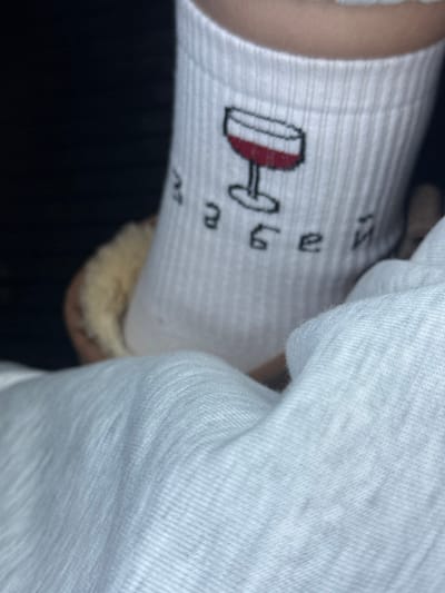 A close-up image displays a lower leg or foot, partially obscured, wearing a white ribbed sock. The sock features a graphic of a wine glass filled with red liquid, accompanied by Cyrillic text which reads "ЗАБЕЙ". The foot appears to be resting in a light-colored, possibly faux-fur lined slipper, partially visible behind the sock. The background is dark and out of focus, suggesting an indoor setting with limited illumination. A white, textured fabric, likely clothing or bedding, occupies the lower foreground. The specific location within Achkhoti, Georgia, cannot be determined from the visual information provided.