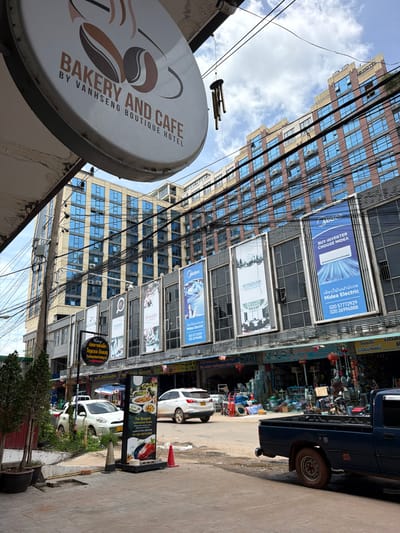 The image shows a street scene in Vientiane Capital, Laos. In the foreground, a sign for "BAKERY AND CAFE BY VANHSENG BOUTIQUE HOTEL" is visible. To the right, a dark blue pickup truck is parked on the side of the road. Further down the street, several buildings line the road, with large advertisements displayed on their facades. These advertisements include brands like "Midea," "Wyndham Hotels & Resort," and "Ramada." The buildings have modern architecture with numerous windows. Various other signs and shop fronts are also visible, suggesting a commercial area. Several cars are parked or driving on the street. The sky is cloudy.
