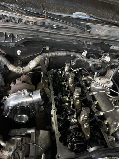 The image displays the open engine bay of a vehicle undergoing maintenance or repair in La Peña, Panama. A new or reconditioned silver turbocharger unit is present on the left side of the engine bay. The engine's cylinder head is exposed on the right, revealing oily camshafts, multiple rocker arms, and fuel injectors with their connected lines, consistent with a diesel engine. Various wiring harnesses, insulated hoses, and metal piping are routed throughout the engine compartment. On the vehicle's cowl near the windshield, a black smartphone, a silver wrench, and a utility knife are visible. The scene indicates an active automotive mechanical operation.