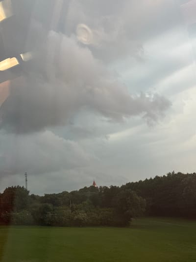 The image shows a landscape scene with a cloudy sky, a tree line, and a grassy field in the foreground. In the distance, on top of a hill covered with trees, there is a structure that appears to be a tower or a building with a red-tiled roof. There is also a tall antenna tower visible on the left side of the hill. The clouds are dark and heavy, suggesting that it might be raining or about to rain. The overall impression is one of a natural landscape under a dramatic sky. The image appears to be taken from a slightly elevated position, looking out over the landscape.