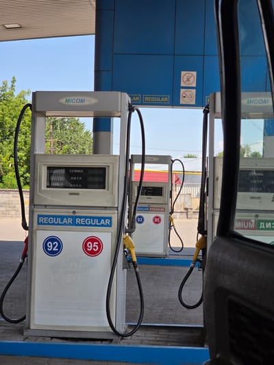 The image shows a gas station with multiple pumps under a covered canopy. The pumps are white and grey with blue trim and displays that show "СУММА" (sum), "ЛИТР" (liter), and "ЦЕНА" (price). Several hoses with red and yellow nozzles are attached to the pumps. The pumps are labeled with "REGULAR" and numbers "92" and "95" inside circles. In the background, there are trees, buildings, and a clear blue sky. There are also two circular signs on the blue wall of the canopy; one shows a crossed-out mobile phone and the other shows a crossed-out cigarette. A portion of a vehicle's interior, including a black door panel and window frame, is visible in the foreground on the right side of the image. The scene appears to be a typical fuel dispensing area at a gas station.