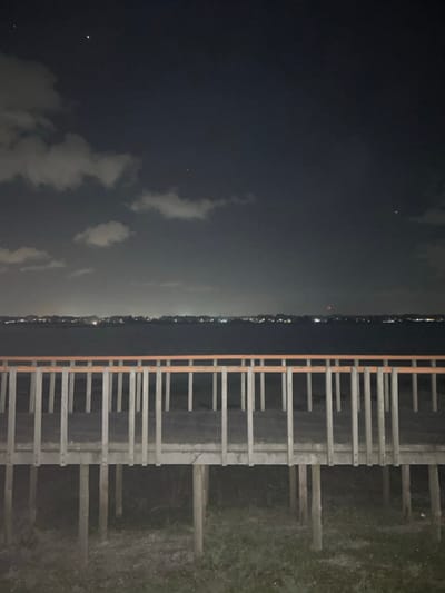 A dark landscape at night showcases a pier or dock on the right, its wooden structure in stark contrast to the darkness. To the left, a body of water, possibly a lake or sea, reflects faint light from a distant shoreline dotted with city lights. Scattered clouds are visible in the night sky, adding texture to the somber scene, and a few stars dot the horizon.