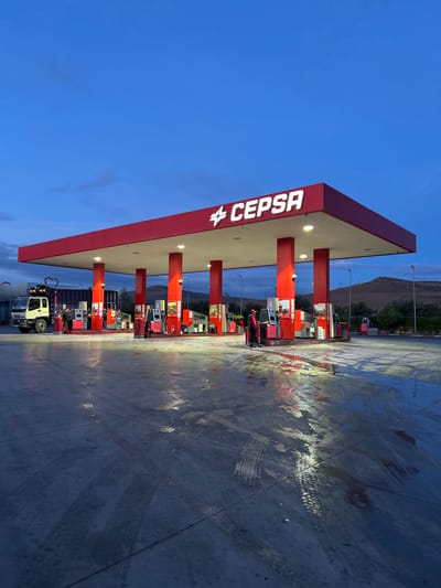 The image depicts a Cepsa gas station at dusk or early night, located in Ain Kansra, Morocco. The scene is dominated by a red and white gas station canopy, illuminated by bright overhead lights. The "CEPSA" logo is prominently displayed in white letters on the red fascia of the canopy.
Beneath the canopy, several red gas pumps are visible, also illuminated. On the ground, the concrete surface appears wet in places, reflecting the artificial lights and showing numerous tire tracks. A white truck is parked under the canopy on the left side of the frame, and a person is visible near one of the pumps, suggesting activity related to refueling.
The sky is a deep twilight blue, indicating either early morning or late evening. In the background, dark hills or mountains are silhouetted against the fading light. The overall impression is of a functional service station operating during twilight hours, with a clear sky above.