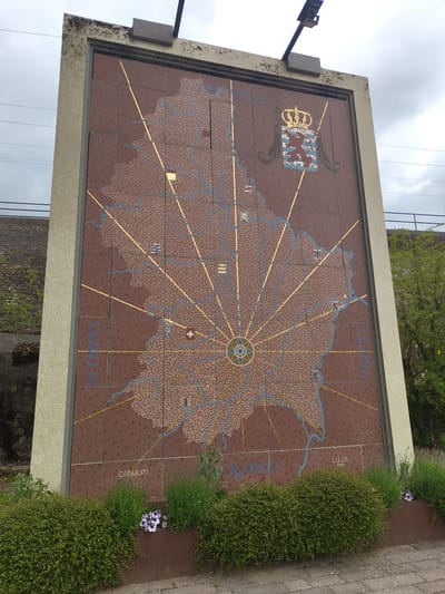 The image shows a large mosaic tile mural. The mural depicts a map of Luxembourg. Prominent features include the country's coat of arms in the upper-left corner, golden lines radiating from a central point, and blue lines indicating rivers and waterways. City names like "ORIUNA" and "LUUA" are visible on the map. The mural is framed by small, off-white tiles. Vegetation, including flowering plants, grows along the edge of the mural. The mural appears to be located outdoors in Wasserbillig, Luxembourg, based on available information.