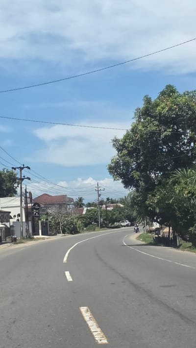 The image depicts a road in Gampongrayadagang, Indonesia. The road curves to the right, with a white line marking the edge. There are intermittent broken white lines in the center of the road. A motorcycle and a car are visible on the road. Trees and buildings line the side of the road. Power lines run along the left side of the road, and the sky is blue with scattered clouds.