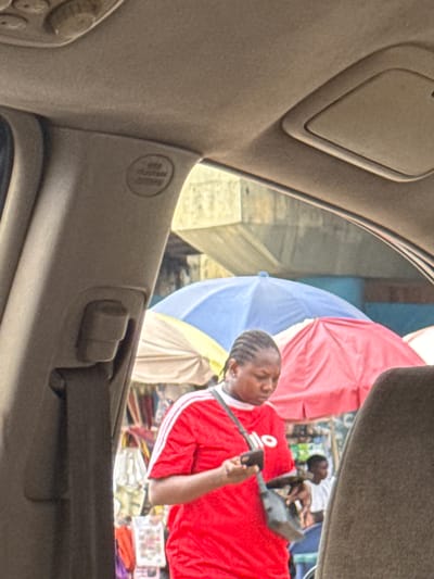 A woman in a red shirt with white stripes on the sleeves is holding a phone and a bag. She is standing in front of stalls covered by umbrellas. The scene appears to be a market in Orji, Nigeria, with several people and goods visible in the background. The image is taken from inside a vehicle, with the car's interior visible in the foreground.