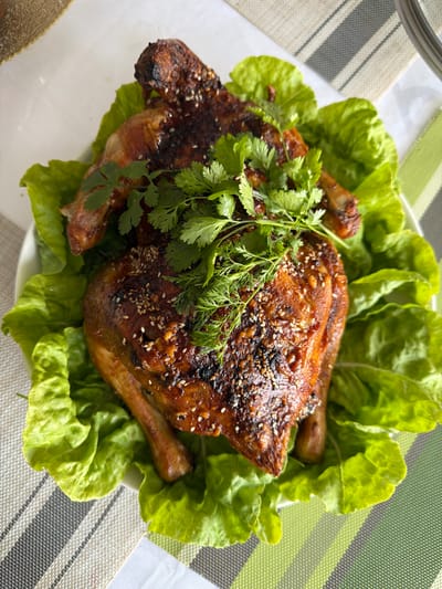 A whole roasted chicken, glistening with a dark glaze and sprinkled with sesame seeds, is presented on a bed of bright green lettuce leaves. Fresh cilantro and dill sprigs are artfully arranged on top of the chicken. The dish is placed on a white plate, which rests on a table covered with a placemat featuring a striped pattern of green, white, and gray. The background is softly blurred, suggesting an indoor setting. This image does not contain any information about the city of Tekeli, Kazakhstan.