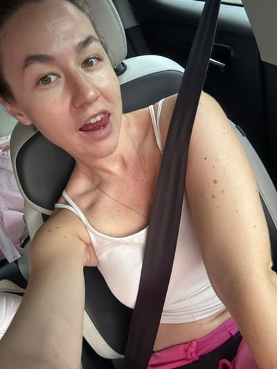 A woman with brown eyes and light skin is sitting in a car, looking at the camera with her tongue sticking out slightly. She is wearing a white tank top and pink shorts, with a black seatbelt across her chest. The interior of the car is visible, with black and white seats. The background is blurry, but appears to be outdoors. There are no other people or animals in the image. The image was taken in Ban Thung Aok, Thailand.