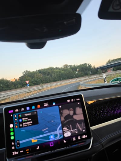 The image shows a view from inside a vehicle driving on a highway near Zeppelinheim, Germany. The dashboard displays a navigation screen with a map and a list of destinations, as well as a music player showing album art and controls. Outside the car, a highway with trees and an overpass is visible under a clear sky. The temperature inside the car is displayed as 23°C.