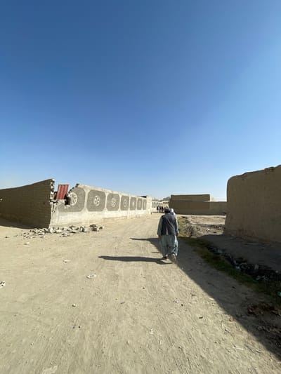 The image depicts an unpaved, dusty road in Spin Boldak, Afghanistan, under a clear, bright blue sky. Harsh sunlight casts long shadows towards the viewer, indicating midday or early afternoon.
On the left side of the road, a partially collapsed brick wall with visible light-colored masonry is present in the foreground, revealing a red corrugated metal sheet behind it, along with scattered debris at its base. Extending further down from this structure is a longer, intact wall with a lighter, possibly concrete, finish on its upper section. This wall features eight evenly spaced, circular decorative motifs, each containing a central cruciform design.
On the right side, an earthen-toned, textured wall or building, likely constructed from mud-brick or adobe, occupies the foreground. A narrow strip of sparse green vegetation and visible refuse runs alongside this wall at the road's edge.
A male figure, viewed from behind, walks away from the camera along the unpaved road, slightly right of center. He is dressed in light blue or grey traditional Afghan attire, including a shalwar kameez and a dark vest. His distinct shadow extends across the road. Further down the road, a small group of at least three additional male figures, also wearing traditional clothing, walk in the same direction.
In the mid-ground, beyond the group of walkers, several low-rise, rectangular buildings constructed from similar materials are visible. A distant, slender structure, possibly a minaret or tower, is faintly visible on the horizon to the left of the road's center. The ground shows tire tracks and scattered minor debris.