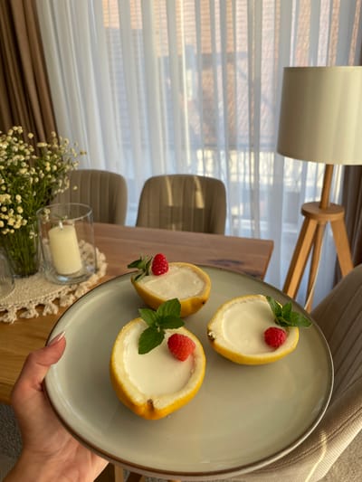 In a close-up, top-down shot, a hand holds a light gray plate featuring a dessert served in hollowed-out lemons. The plate contains two halves of a lemon, each filled with a creamy white dessert, garnished with a fresh raspberry and a sprig of mint. The scene is set indoors, likely a home, with soft, diffused light suggesting daytime. In the background, a wooden lamp and striped blinds are visible, creating a warm and inviting atmosphere. A bouquet of small white flowers and a lit candle in a glass holder are placed on a wooden surface in the foreground, adding to the cozy ambiance. The overall impression is one of a carefully prepared and aesthetically pleasing dessert, presented in a natural and elegant manner. There is no visible text in the image.