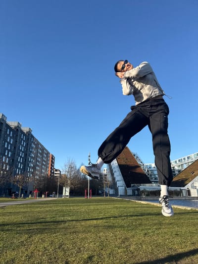 A person is captured performing a backflip in a park-like setting in Tårnby, Denmark. They are wearing a light-colored hooded sweatshirt, black sweatpants, white socks, and sneakers. The backdrop includes a blue sky, green grass, buildings of varied architectural styles, and bare trees. The environment suggests an urban park or recreational area.