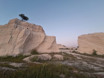 This media file depicts an outdoor scene in Fetești, Moldova. The primary subjects are a sandy, hilly landscape with a clear sky. Dominating the left side of the image is a large, light-colored rock formation with visible erosion patterns. A lone tree with dark foliage is perched precariously on the highest point of this formation. In the mid-ground, a smaller, rounded rock formation stands to the right, also appearing eroded. The foreground is covered in dry grass and sparse yellow wildflowers. The ground itself appears sandy and uneven, with tire tracks visible in some areas, suggesting a path or track.
The time of day appears to be either early morning or late evening, indicated by the soft, diffused light and the clear, pale blue sky transitioning to a faint pinkish hue near the horizon. The weather seems clear and calm. There is no discernible activity or event taking place; the scene is still and natural. No people, animals, or man-made objects, other than the implied tracks, are visible. The overall mood is one of serene, rugged natural beauty. No text is visible in the image.