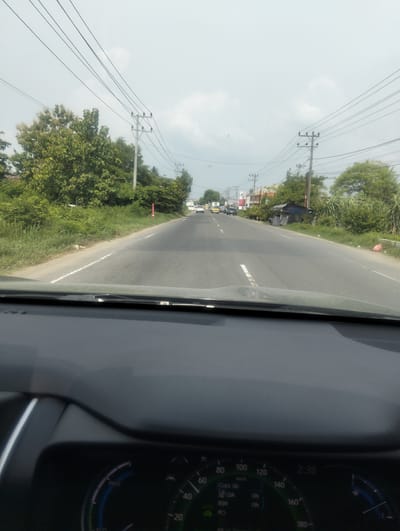 The image depicts a forward-facing view from the interior of a vehicle driving on a two-lane asphalt road. The road is flanked by lush green vegetation and trees. Utility poles with multiple wires are visible along the side of the road. In the distance, there are buildings and other vehicles, including a truck and a few cars. The sky is overcast with a pale, hazy appearance. The dashboard of the vehicle is partially visible in the foreground, displaying a speedometer and other indicators. The dashboard shows the time as 2:38 and various labels such as "PWR", "ECO", "LDA", "BSM", and "PCS". The setting appears to be a semi-rural or suburban area on a cloudy day.