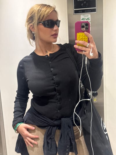 An individual is depicted taking a mirror selfie in what appears to be an elevator, located in an unidentified building within Nogent-sur-Marne, France. The subject, appearing to be a young adult female with light-colored hair styled in an updo, wears dark wraparound sunglasses, small silver earrings, a thin necklace, and a small neck tattoo. She is dressed in a black long-sleeved, button-front ribbed top. A dark, possibly black or navy, long-sleeved garment is tied around her waist, and light brown or beige ribbed fabric is visible below. Her left wrist displays stacked green bracelets, and her left hand, positioned on her hip, has multiple rings on several fingers. She holds a purple-cased smartphone, equipped with a yellow textured grip accessory and white wired earphones, in her right hand. A black bag with faint white markings is slung over her left shoulder. The background reflection consists of silver-colored metal panels featuring elevator control buttons, including an 'up' arrow and a door-open symbol.