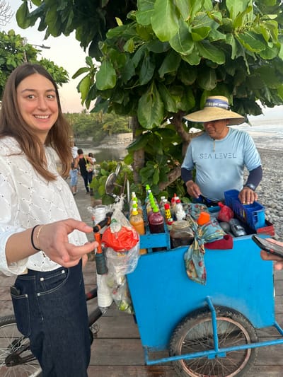 A woman smiles, facing the camera, with dark jeans and a white blouse. Behind her is a man with a straw hat, preparing drinks from a blue cart with various colorful liquids. The cart has a single wheel. Lush green trees frame the scene, and a beach and blurred figures are visible in the background.