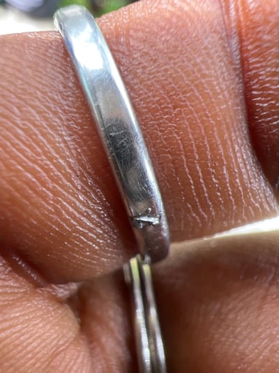 The media file is a close-up, macro photograph of a person's hand. The focus is on a silver ring that appears to be slightly damaged, with a small nick or crack visible on its surface. The skin of the hand is detailed, showing the natural lines and texture. The background is out of focus, suggesting a natural setting with some greenery visible, though not clearly identifiable. The lighting appears to be natural daylight, casting highlights on the ring and the skin. There is no visible text in the image. The image captures a moment of close examination of the ring on the hand. Given the location context of Yali, Nigeria, the skin tone suggests the person is likely of African descent. The overall impression is one of detail and texture, highlighting the intricate surface of both the human skin and the metallic object.