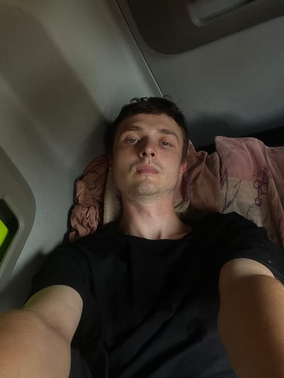 The image shows a close-up, high-angle, selfie-style shot of a young man lying down. He is wearing a black t-shirt and appears to be on a bed or soft surface, as indicated by a pink and patterned cloth underneath his head. The background is a muted gray, with some texture suggesting a padded or upholstered surface. The lighting appears to be artificial, and it's difficult to determine the time of day or weather.

The man's face is the primary focus. He has short, dark hair and light-colored eyes. His expression is neutral, with his gaze directed towards the camera. There is some stubble on his chin and jawline. His right arm is visible, bent at the elbow and resting on the fabric.

The scene appears to be indoors, possibly in a room with neutral-colored walls or furnishings. The gray surface could be part of a bed, a couch, or even a vehicle interior. No text is visible in the image. The overall mood is relaxed or perhaps introspective, given the man's pose and direct gaze. The setting appears to be domestic or a similar private space.