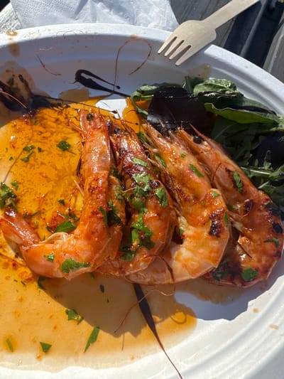 The image depicts a plate of grilled shrimp served with greens and a wooden fork. The shrimp are arranged in a stack, coated with what appears to be a glaze or sauce and sprinkled with herbs. The greens include arugula and what might be beet greens. The plate is white and appears to be made of paper. The setting seems to be outdoors, possibly on a picnic table, as suggested by the wooden surface visible at the left. There is no clear evidence to confirm that it is specifically in Epping Forest, United Kingdom.